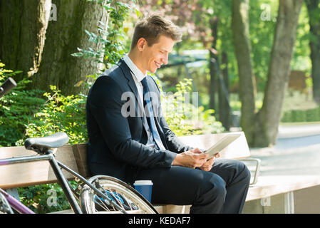 Male businessman sitting on bench using digital tablet at park Banque D'Images