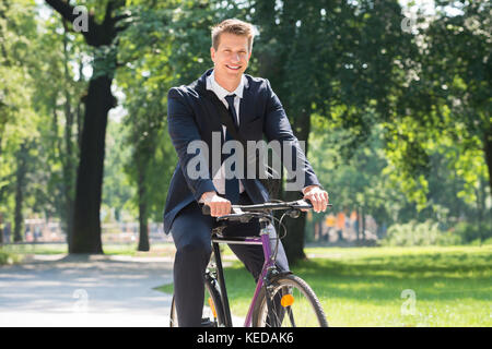 Heureux jeune homme businessman riding bicycle in park Banque D'Images