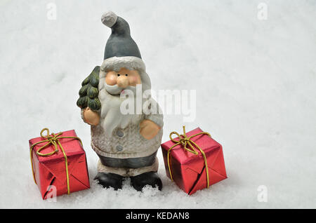 Figurine en céramique de Père Noël avec des vêtements gris, un arbre de Noël et deux parcelles rouges sur fond de neige, horizontale Banque D'Images