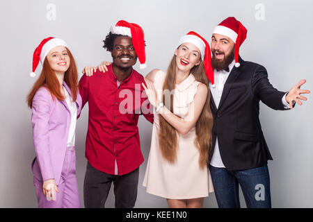 Réunion d'amis pour célébrer Noël. wearing santa bonnet rouge, rugueux et souriant à la caméra à l'intérieur. plan, arrière-plan gris Banque D'Images