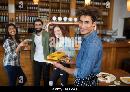 Portrait of smiling waiter serving tray de mini-maïs à des clients au bar Banque D'Images