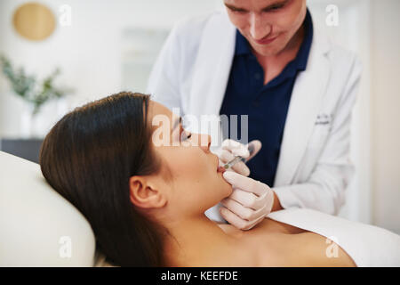 Jeune homme médecin d'effectuer des injections de botox pour les lèvres d'une femme brune allongé sur une table dans une clinique de beauté Banque D'Images