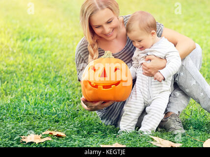 Mère heureuse avec un petit bébé à l'extérieur, assis sur la pelouse verte et orange citrouille sculptée avec jouer avec visage souriant,bénéficiant d'halloween h Banque D'Images