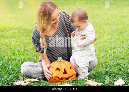 Famille heureuse célébrer Halloween en plein air, mère avec mignon bébé Garçon jouant avec la citrouille sculptée sur le champ vert, profitant de l'automne maison de vacances Banque D'Images