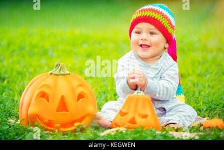 Cute little baby boy wearing colorful funny hat sitting in backyard on Green grass prairie près de deux citrouilles, jeux en plein air avec halloween traditionnelle Banque D'Images