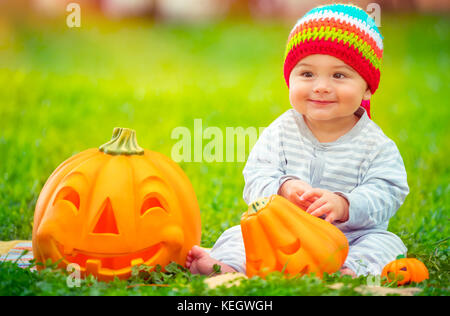 Cute little baby boy wearing colorful funny hat, assis dans la cour sur l'herbe verte prairie près de deux citrouilles décoratives, jeux en plein air avec traditi Banque D'Images