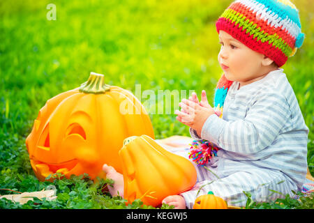 Mignon petit garçon assis sur l'herbe verte domaine portant chapeau coloré et amusant de jouer avec les citrouilles avec smiling visages sculptés, happy Halloween de Banque D'Images