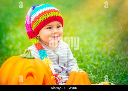 Joli petit enfant assis sur l'herbe verte champ près de deux citrouilles mignon avec visages sculptés, jouant avec décoration de fête, célébrer Halloween holi Banque D'Images