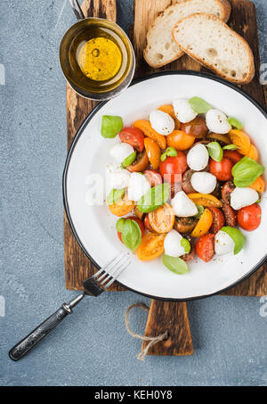 Salade caprese italien avec tomates cerises, mozzarella et basilic frais petits en émail blanc sur plaque en bois rustique au béton gris backgroun Banque D'Images