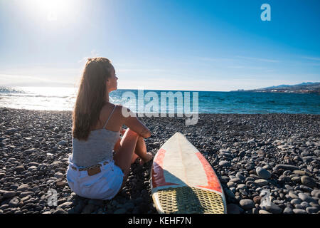 Femme caucasienne assise sur la plage près de la planche de surf Banque D'Images