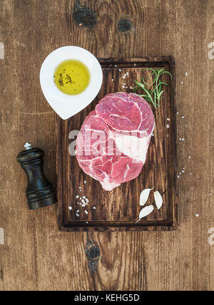 La viande fraîche de boeuf cru cross cut pour couper ossobuco avec gousses d'ail, le romarin, le poivre, l'huile et le sel sur le service à bord sur fond de bois rustique, haut Banque D'Images