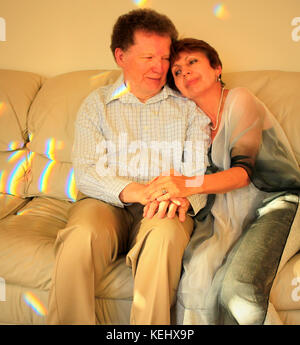 Vieux couple assis sur un canapé éclairée par la lumière du soleil se tenant la main ; la femme repose sa tête sur l'épaule de l'homme tandis que l'homme est à la recherche de son sourire. Banque D'Images