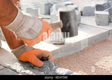 Les mains dans les gants d'un travailleur constructeur pose sur les pavés de granit sett avec marteau maillet en caoutchouc et le pavage des trottoirs de la rue on construction site Banque D'Images