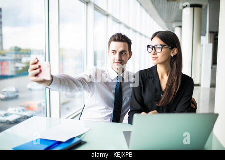 Deux collègues amis de prendre la photo d'eux-même assis dans le bureau, l'homme et de la femme en tenant l'appareil photo avec téléphone selfies, drôle de travail dans le Banque D'Images