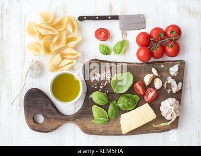 Ingrédients pour la cuisson des pâtes. conchiglioni, feuilles de basilic, tomates cerise, parmesan, huile d'olive, sel, ail sur planche à découper en noyer rustique sur fond de bois blanc Banque D'Images