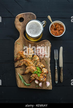 Ailes de poulet frit sur du conseil rustique, épicé de la sauce tomate, les herbes et mug de bière légère sur toile en bois noir Banque D'Images