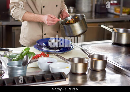 Chef publié avec des légumes à l'étouffée cuillère. Portrait of male chef va mettre la garniture en acier inoxydable bol. la préparation de l'aliment à la professional Banque D'Images
