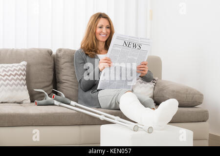 Femme mature avec fracture de la jambe reading newspaper in house Banque D'Images