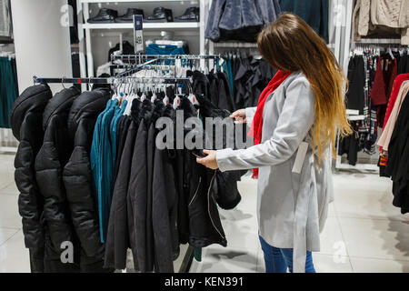 Shopping, la mode, la vente, la ,style et personnes concept - jeune femme en manteau gris et foulard rouge choisir veste noire dans une boutique de vêtements. Banque D'Images