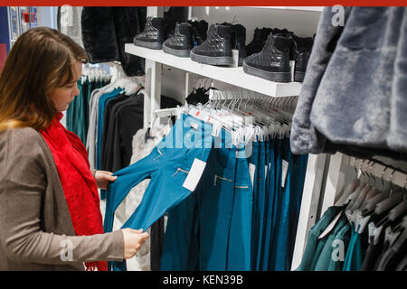 Shopping, la mode, la vente, la ,style et personnes concept - jeune femme en manteau gris et foulard rouge choisir blue trousers in clothing store Banque D'Images