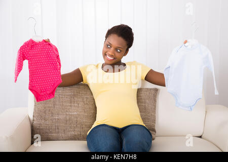 Happy pregnant woman Holding Baby Clothes, assis sur un canapé à la maison Banque D'Images