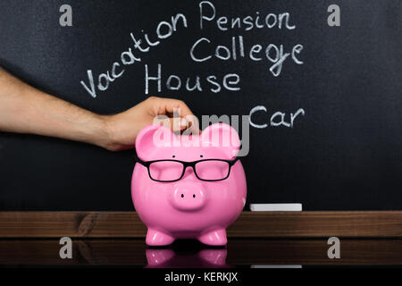 Personne hand inserting coin dans la tirelire pour les dépenses quotidiennes in front of blackboard Banque D'Images