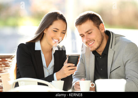 Deux cadres heureux d'avoir l'oeil sur le contenu en ligne dans un téléphone mobile assis dans un restaurant terrasse Banque D'Images