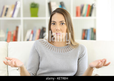 Vue de face d'une femme en haussant les épaules et douteux en vous regardant assis sur un canapé à la maison Banque D'Images