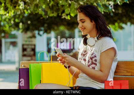 Young beautiful smiling girl, un adolescent, en appui sur le banc après le shopping. à côté d'elle sont des sacs pleins, choses qu'elle a acheté. Elle est titulaire d'un téléphone mobile Banque D'Images