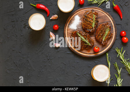 Des steaks délicieux barbecue et de la bière dans des verres sur une pierre noire, avec des tomates cerises et des piments, romarin. foi Banque D'Images
