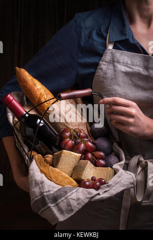 Femme tenant un panier avec des fromages, vin, fruits et pain. Dans la composition des couleurs sombres Banque D'Images