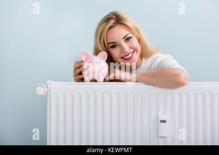 Jeune femme heureuse avec tirelire sur radiateur à la maison Banque D'Images