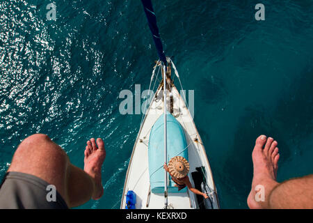 Un homme se penche contre le gréement vu d'en haut et encadré par les pieds d'un homme sur le mât après avoir enveloppé la voile au large de Glovers Reef sur la côte de Belize. Banque D'Images