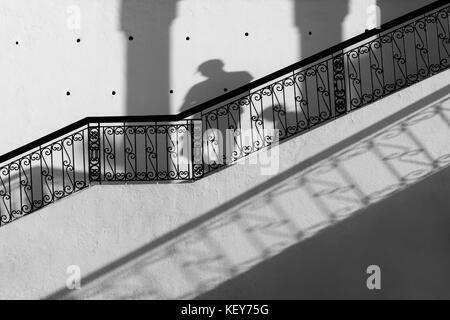 Escalier avec rampe en noir contre mur blanc, avec les ombres d'un homme marchant vers le bas. noir et blanc, image monochrome. Banque D'Images