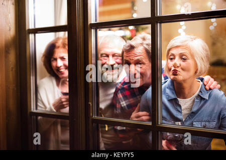 Amis seniors avec des verres à vin à Noël. Hommes et femmes regardant par la fenêtre, s'amusant. Tiré à travers le verre. Banque D'Images
