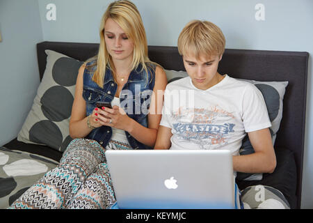 Amis adolescents, assis sur un lit, garçon en utilisant son ordinateur portable tandis que sa petite amie est occupé avec son téléphone intelligent Banque D'Images