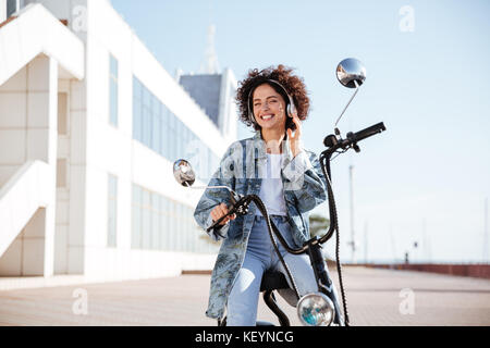 Smiling curly girl moto moderne à l'extérieur et à l'écoute de la musique Banque D'Images