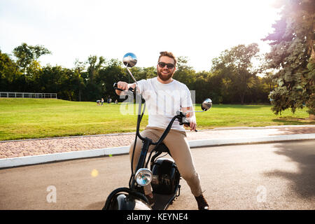 Heureux homme barbu à lunettes assis sur moto moderne à l'extérieur et à l'appareil photo Banque D'Images