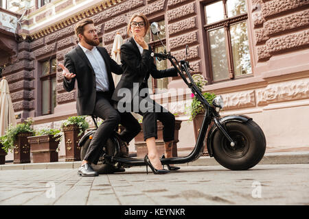 Vue latérale sur toute la longueur de l'image déplut business man sitting on barbu moto moderne en plein air avec des jeunes femme élégante qui à miroir à Banque D'Images