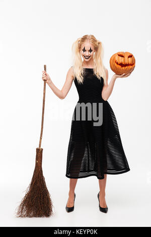 Portrait of a smiling woman fou habillé en costume debout avec un balai et Halloween Pumpkin isolated over white background Banque D'Images