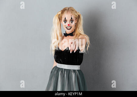 Portrait of a young blonde woman in clown halloween make-up posing while standing et pointant sur fond gris plus isolé de l'appareil photo Banque D'Images