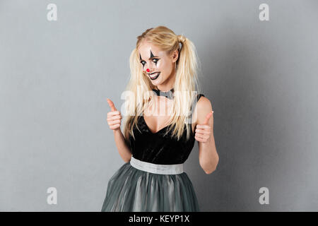 Portrait of a young blonde woman in clown Halloween maquillage showing Thumbs up geste en se tenant debout et looking at camera isolated over gray Banque D'Images