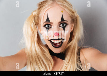 Portrait d'une femme blonde excité en clown halloween make-up en prenant un isolé sur fond gris selfies Banque D'Images