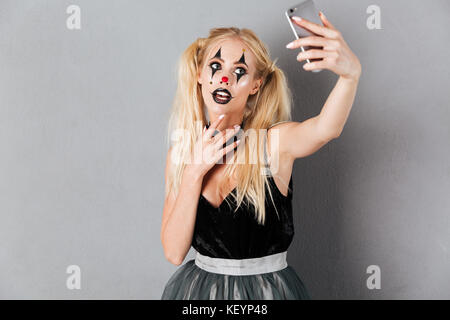 Portrait d'une jeune femme blonde en clown halloween make-up en tenant un téléphone mobile avec selfies et posant sur fond gris isolé Banque D'Images