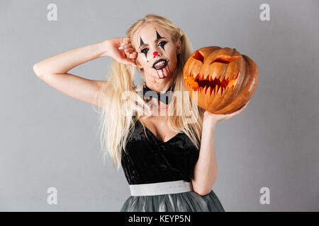 Jeune femme blonde dans halloween composent posant avec citrouille sculptée et regardant la caméra sur fond gris Banque D'Images