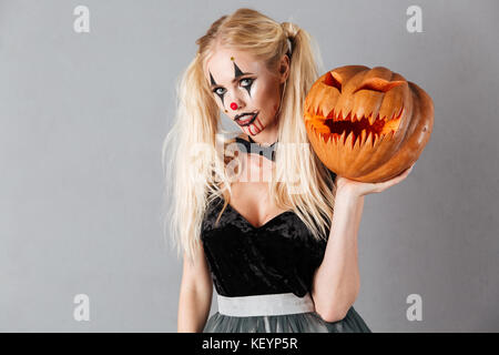 Jeune femme blonde en clown Halloween maquillage et des traînées de sang et à la citrouille - tenue à huis clos sur fond gris isolé Banque D'Images