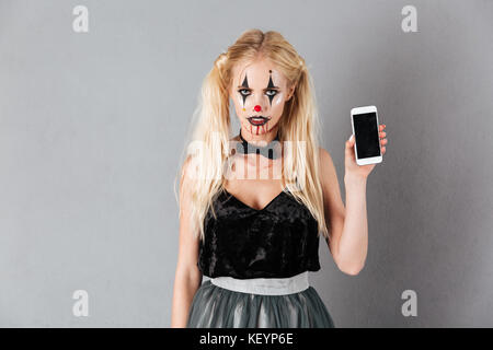 Portrait of a young blonde woman in Halloween maquillage de clown montrant l'écran vide isolé téléphone mobile sur fond gris Banque D'Images