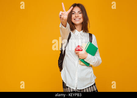 Portrait of a cheerful pretty schoolgirl holding books et apple en position debout et montrant la paix isolé sur fond orange geste Banque D'Images