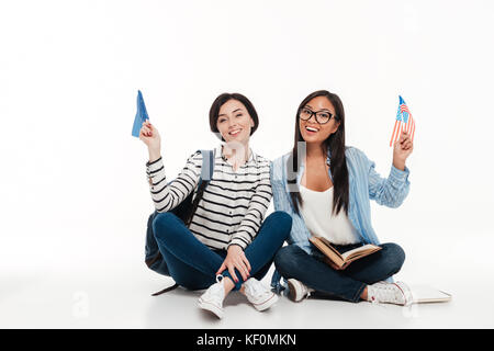 Deux jeunes female students holding drapeaux américains tout en restant assis ensemble sur le plancher isolé sur fond blanc Banque D'Images