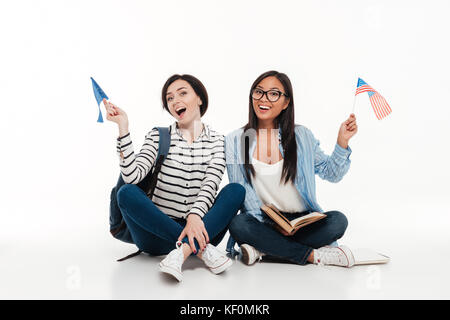 Deux jeunes étudiantes gaies holding drapeaux américains tout en restant assis ensemble sur le plancher isolé sur fond blanc Banque D'Images